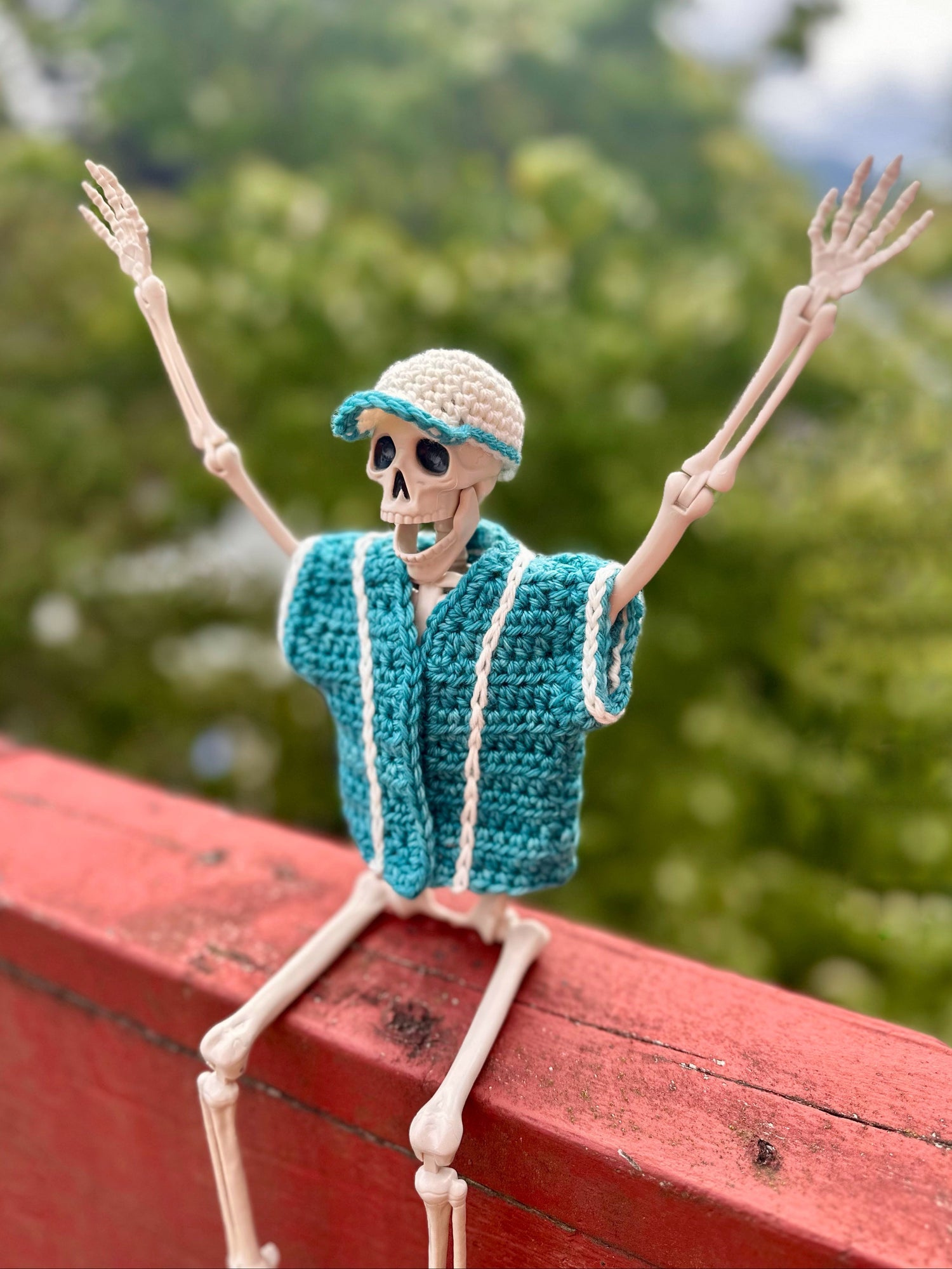 Happy skeleton figure with its arms raised in joy, wearing a blue crochet baseball jersey and hat on a wooden bench with a blurred green background