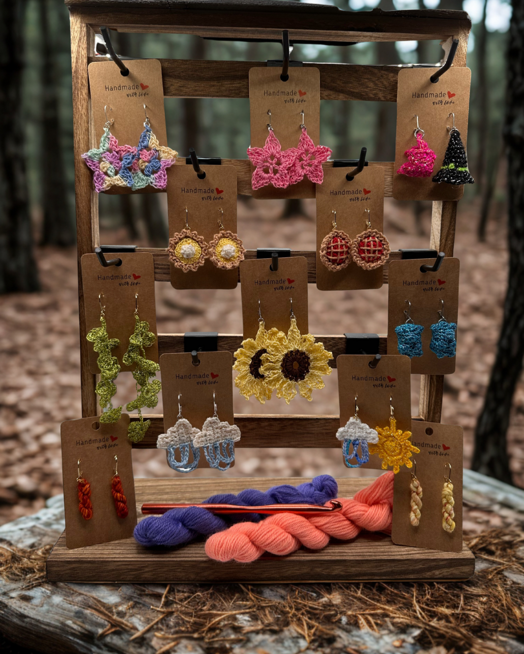 Display of colorful, handmade cotton and yarn earrings on a wooden stand with yarn skeins and a crochet hook in a forest setting.