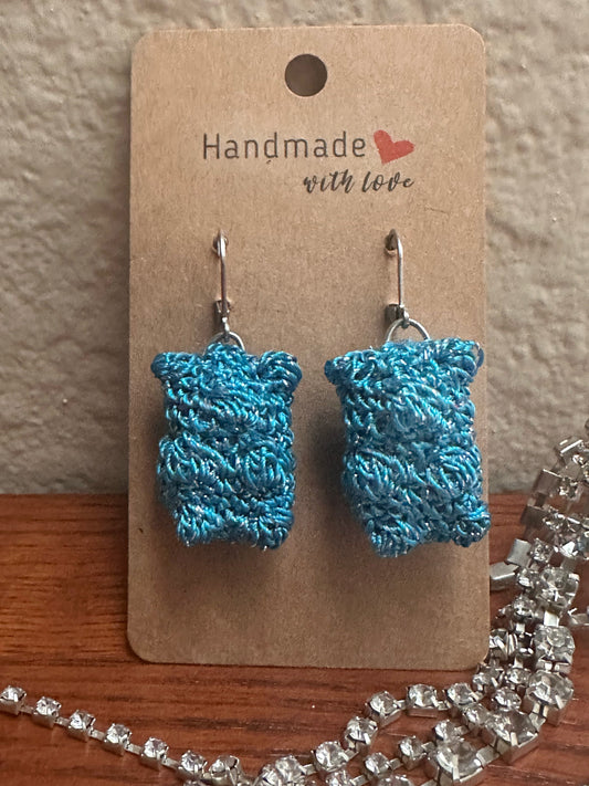 Blue sparkly gummy-bear-shaped earings on a cardboard backing and resting on a wooden shelf with crystals lying around it. 