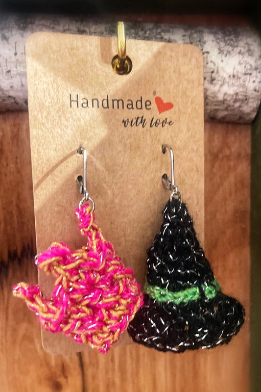 Micro crocheted earrings on a wooden stand. One earring is a black witches hat, the other is a pink crown, both are sparkly and handmade. 